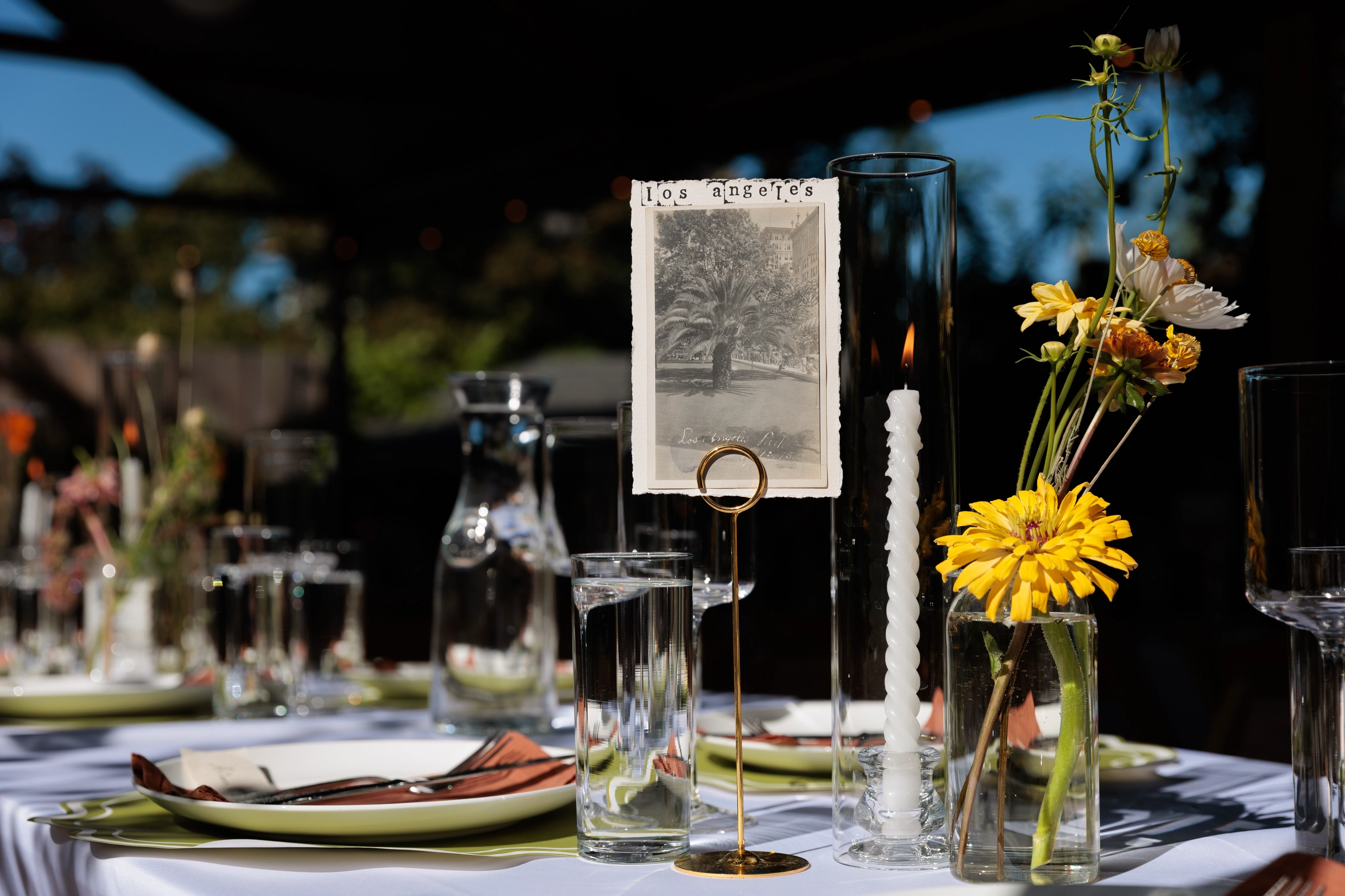 Decor setup of green place settings, lit, white wax candle, Los Angeles postcard, water glass, and yellow wildflowers.